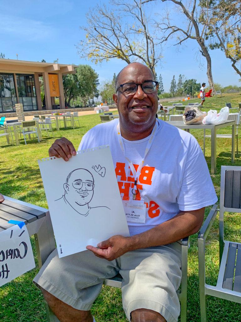 An alum holds up a cartoon drawing of himself while sitting on the mounds. 